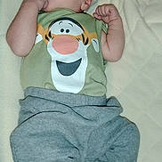 Joseph is registered to the contest to win money with this photo: baby, infant, child, clothing, green_shirt, cartoon_print, gray_pants, blanket, hand, feet, face, lying_down, cute, skin, expression, indoors, soft_texture, portrait, newborn, small