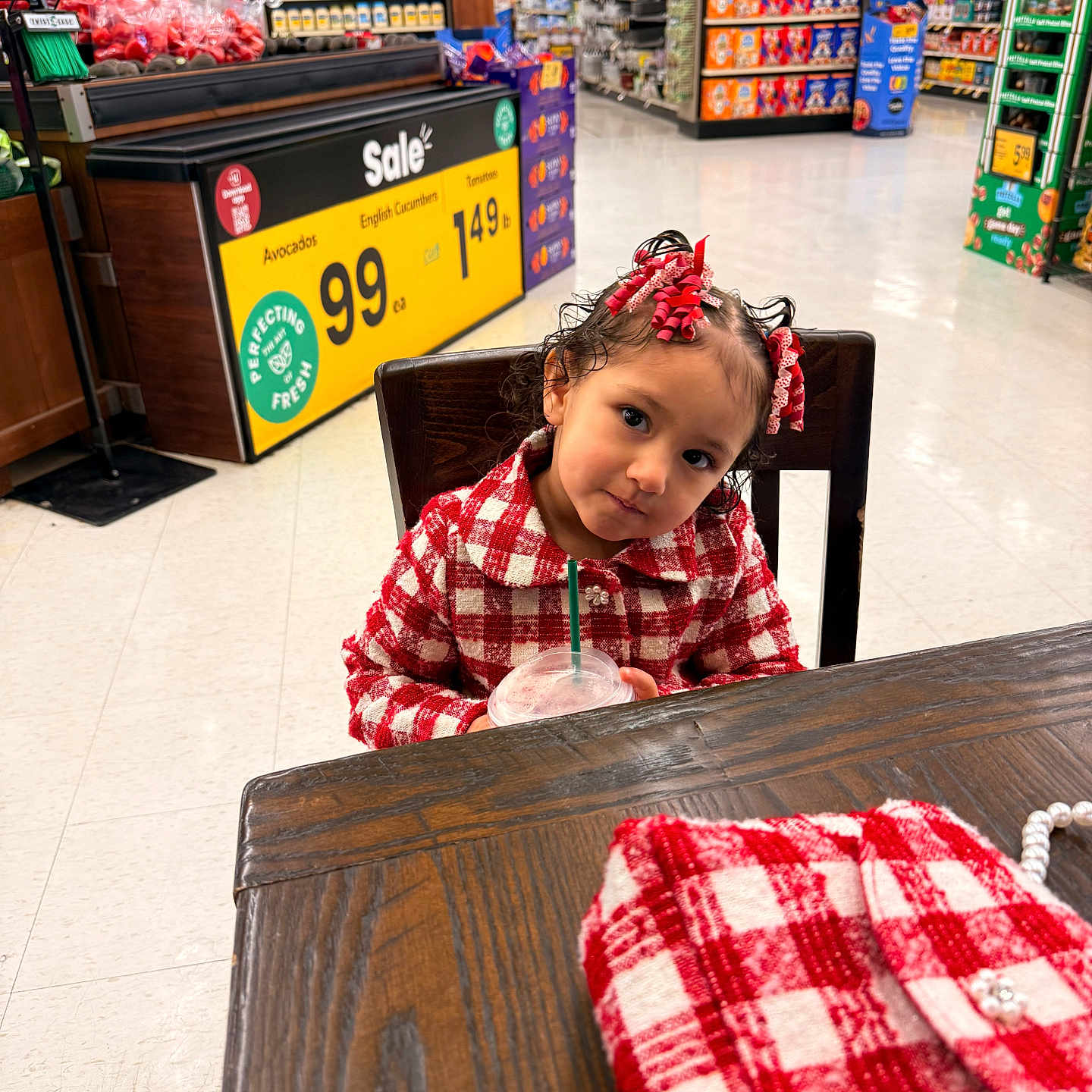 Alaura joined the competition — help win amazing prizes! child, girl, red_checkered_clothing, hair_ribbons, wooden_table, drink, straw, grocery_store, sale_sign, avocados, cucumbers, aisle, shelves, floor, chair, expression, head_tilt, beverage, shopping, indoor