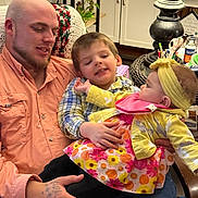 Esther is registered to the contest to win money with this photo: man, child, baby, family, smiling, indoor, living_room, lamp, plant, furniture, colorful_clothing, headband, floral_dress, plaid_shirt, tattoo, holding, happy, seated, cozy, home