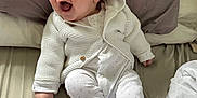 Charlie is registered to the contest to win money with this photo: baby, infant, smile, white_clothing, hoodie, knitted, ear_hood, bed, pillow, blanket, happy, cute, child, footed_pajamas, person, indoors, lying_down, expression, young, cozy