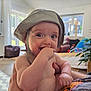 baby, child, hat, hand, indoor, person, skin, face, diaper, furniture, plant, light, window, curtain, couch, arm, smile, living_room, cheek, holding