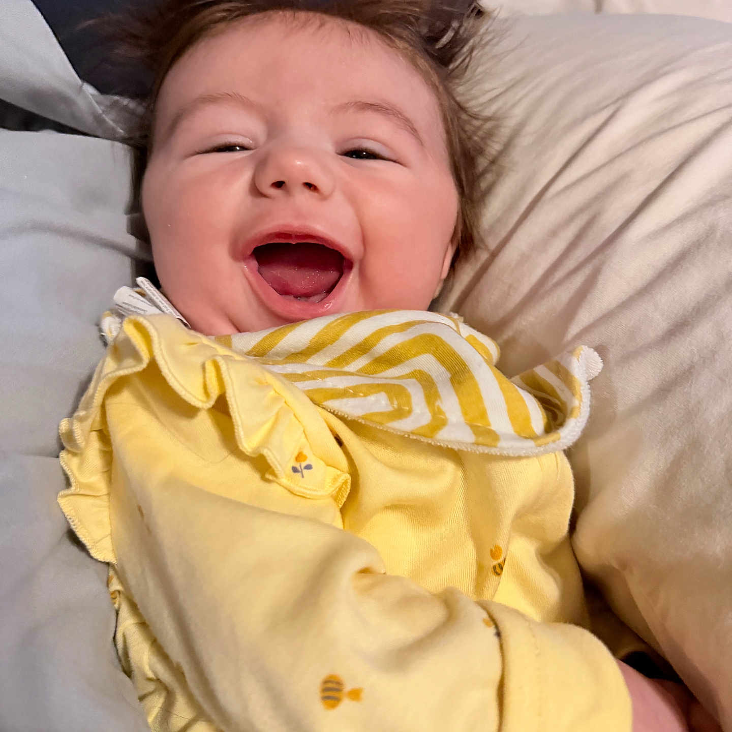 Scarlett is registered to the contest to win money with this photo: baby, smiling, happy, yellow_clothing, pajamas, bed, pillow, face, child, infant, cute, joyful, hair, skin, person, lying_down, indoors, closeup, portrait, sleepwear
