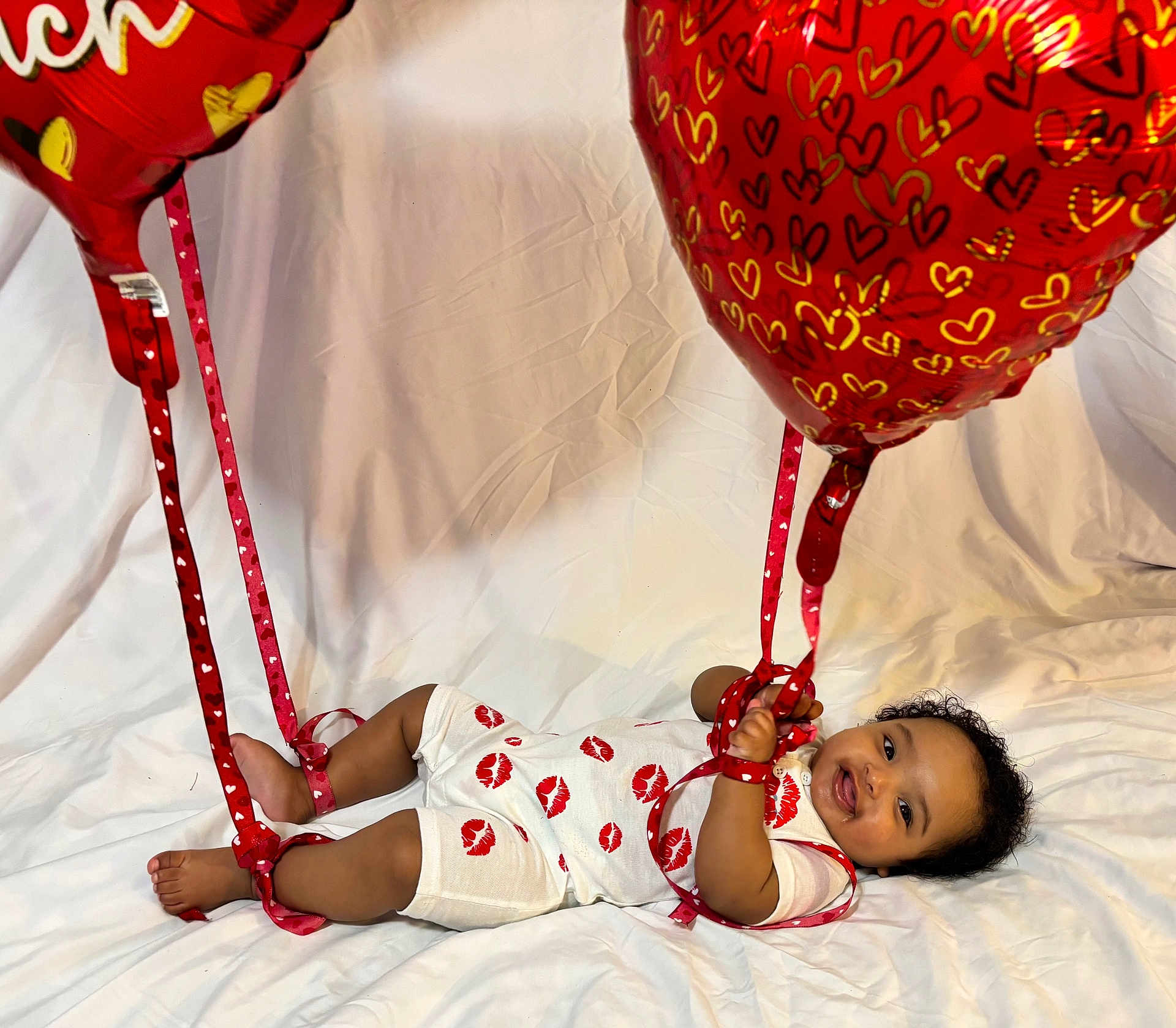 Santi is registered to the contest to win money with this photo: baby, infant, child, balloons, red_balloon, heart_balloon, ribbon, ribbons, kiss_prints, onesie, smile, happy, lying_down, white_sheet, curly_hair, portrait, studio_shot, celebration, cute, hands