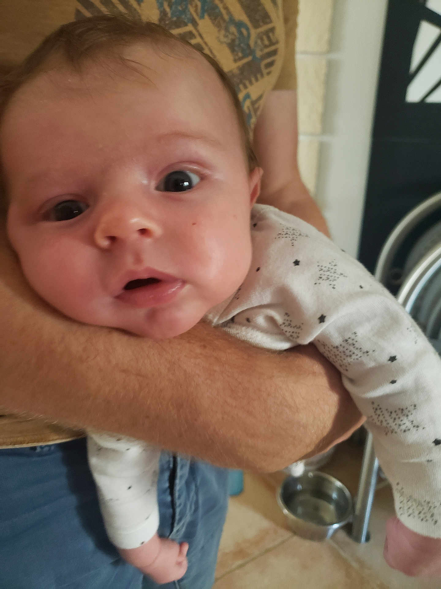 Arthur a rejoint le concours — aidez-le/la à gagner de superbes lots ! baby, infant, person, arm, clothing, white_outfit, stars_pattern, face, eyes, mouth, hand, indoor, floor, tile_floor, pet_bowl, adult, holding, closeup, expression, surprised