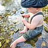 baby, child, water, stream, rocks, hat, blue_cap, suspenders, barefoot, outdoor, nature, curious, sitting, exploring, summer, sunlight, reflection, skin, legs, hands