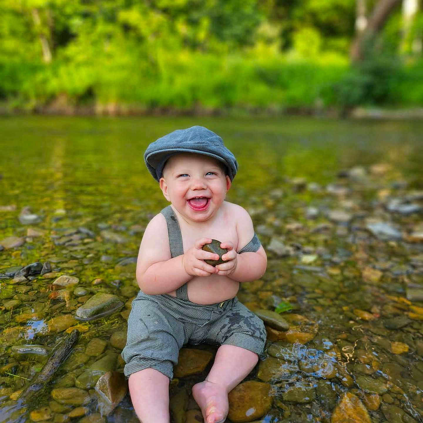 Reid joined the competition — help win amazing prizes! baby, child, water, river, rocks, nature, outdoor, smiling, happy, cap, suspenders, barefoot, sitting, greenery, shallow_water, playful, summer, portrait, cute, sunlight