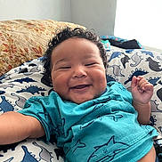 Asiyah is registered to the contest to win money with this photo: baby, smile, curly_hair, dinosaur_pattern, blanket, blue_shirt, indoor, natural_light, happy, infant, cute, child, person, lying_down, cozy, clothing, pattern, window, closeup, skin
