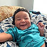 baby, smile, curly_hair, dinosaur_pattern, blanket, blue_shirt, indoor, natural_light, happy, infant, cute, child, person, lying_down, cozy, clothing, pattern, window, closeup, skin