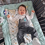 Raphaël participe au concours pour gagner de l'argent avec cette photo : baby, crib, overalls, bib, toy, blanket, smiling, happy, indoor, child, footwear, white_shoes, patterned_blanket, soft_toys, wooden_floor, playpen, infant, cute, lying_down, clothing