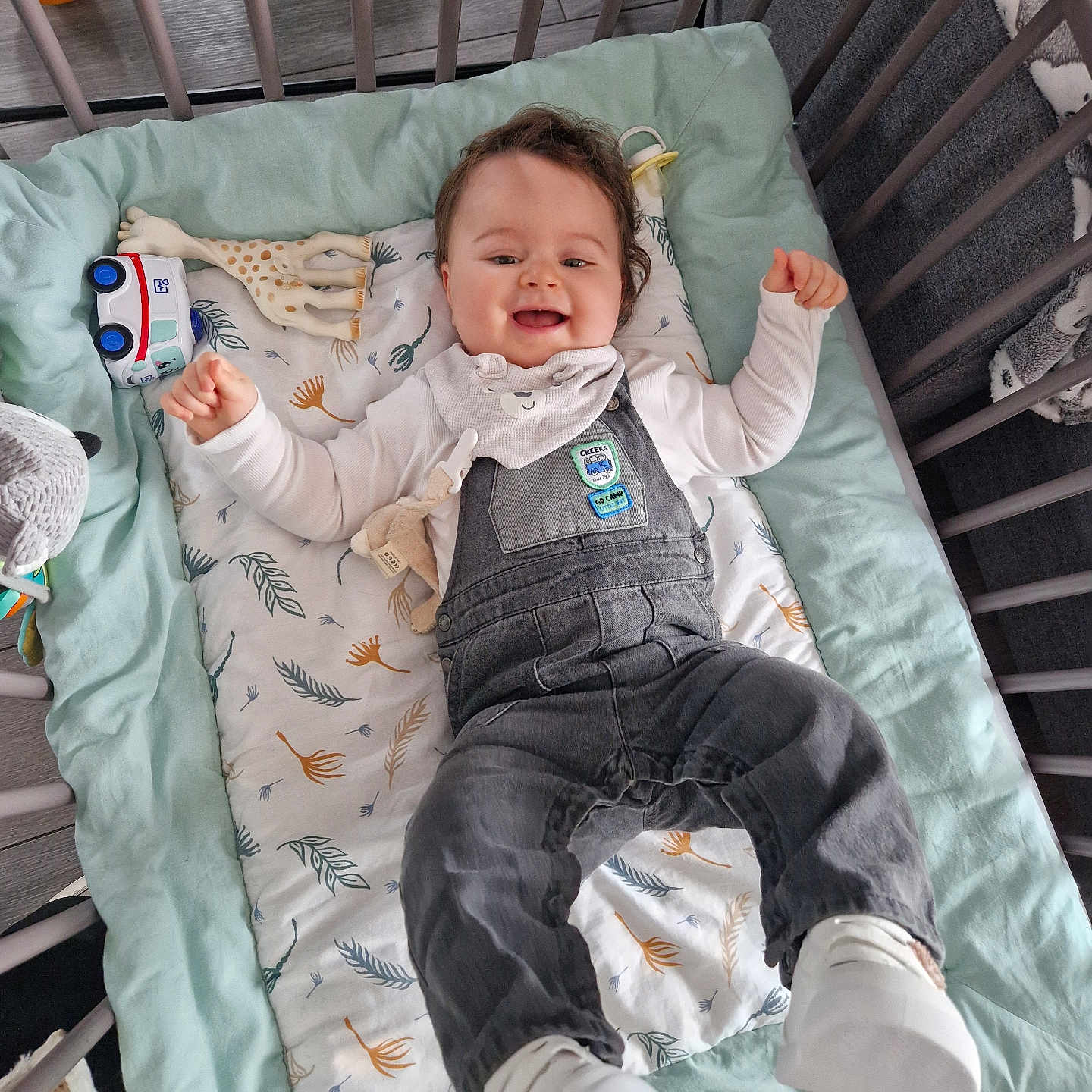 Raphaël participe au concours pour gagner de l'argent avec cette photo : baby, bib, blanket, child, clothing, crib, cute, footwear, happy, indoor, infant, lying_down, overalls, patterned_blanket, playpen, smiling, soft_toys, toy, white_shoes, wooden_floor