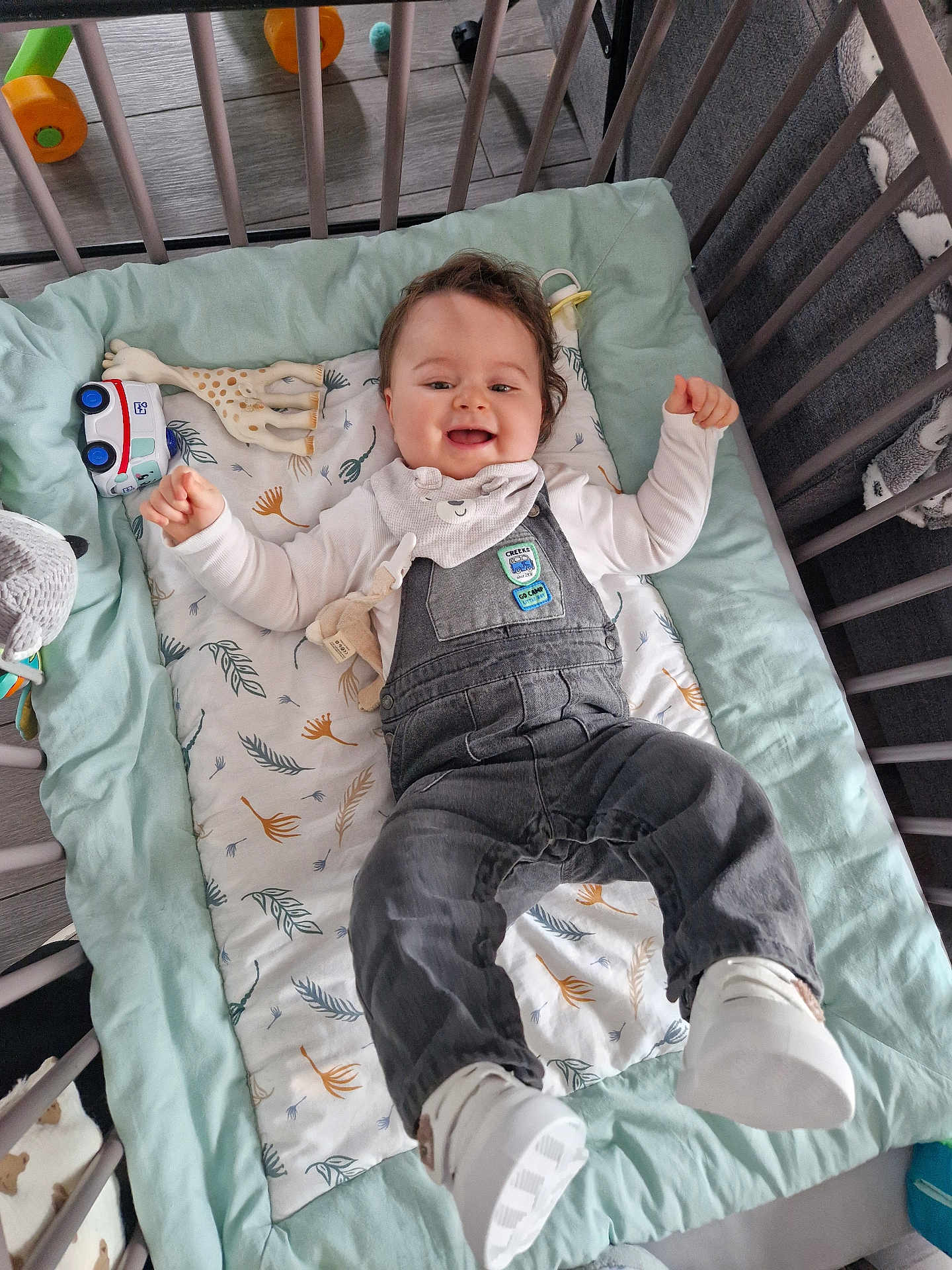 Raphaël participe au concours pour gagner de l'argent avec cette photo : baby, crib, overalls, bib, toy, blanket, smiling, happy, indoor, child, footwear, white_shoes, patterned_blanket, soft_toys, wooden_floor, playpen, infant, cute, lying_down, clothing