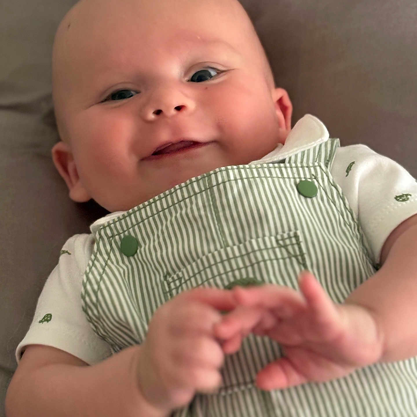 Simon is registered to the contest to win money with this photo: baby, button, cheeks, child, clothing, cute, face, green, hands, happy, infant, lying_down, overalls, person, portrait, skin, smile, soft_background, striped, white_shirt
