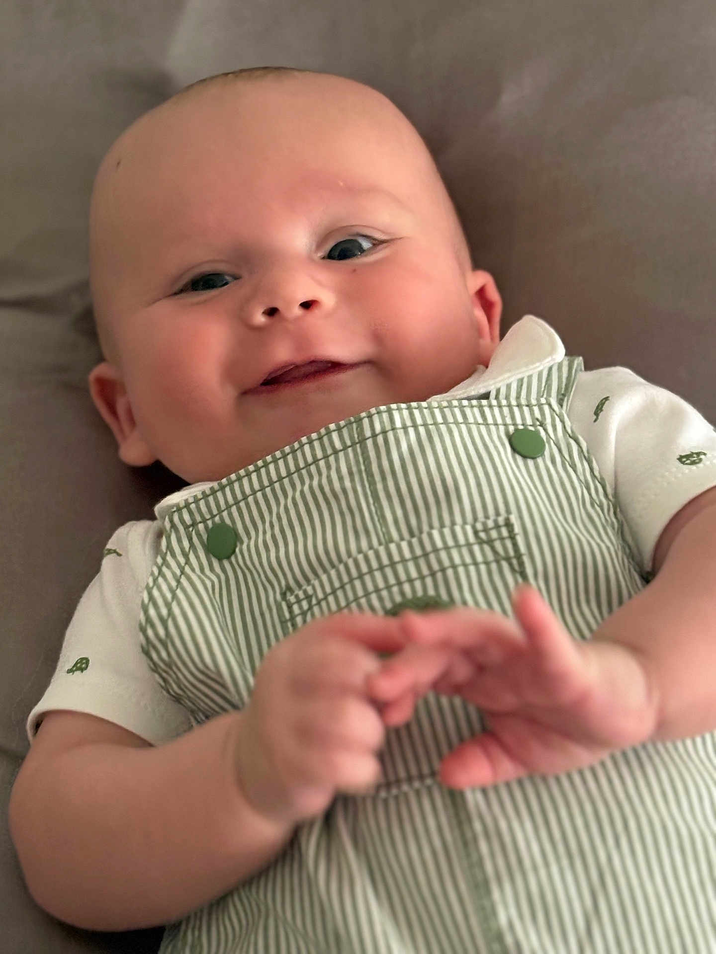 Simon is registered to the contest to win money with this photo: baby, smile, face, hands, overalls, clothing, infant, lying_down, cute, child, portrait, skin, cheeks, white_shirt, button, green, striped, happy, person, soft_background