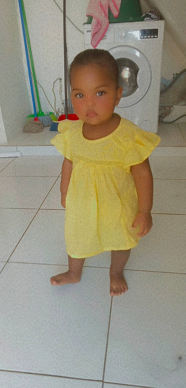 Mayra participe au concours pour gagner de l'argent avec cette photo : arm, baby_toddler_clothing, barefoot, chin, day_dress, eye, face, floor, flooring, head, iris, knee, neck, one_piece_garment, person, skin, sleeve, thumb, toddler, trunk