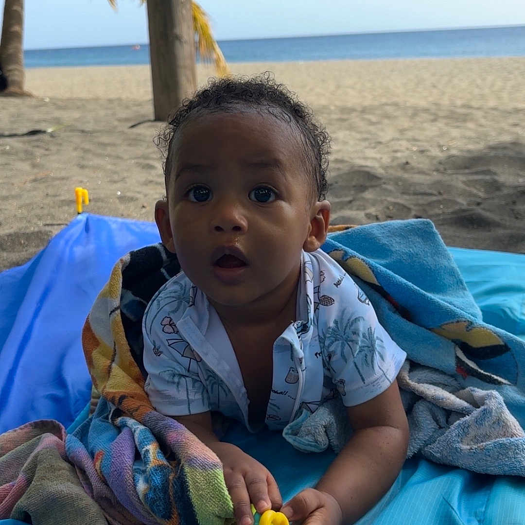 Kaëyson a rejoint le concours — aidez-le/la à gagner de superbes lots ! baby, beach, blanket, blue, child, clothing, curly_hair, cute, infant, nature, ocean, outdoor, palm_tree, playful, portrait, relaxation, sand, summer, toy, tropical