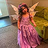 Emelina is registered to the contest to win money with this photo: child, girl, fairy_costume, purple_dress, flower_crown, wings, indoor, carpet, green_couch, wall_art, playful, pouting, curly_hair, portrait, person, costume, dress_up, smile, home, furniture