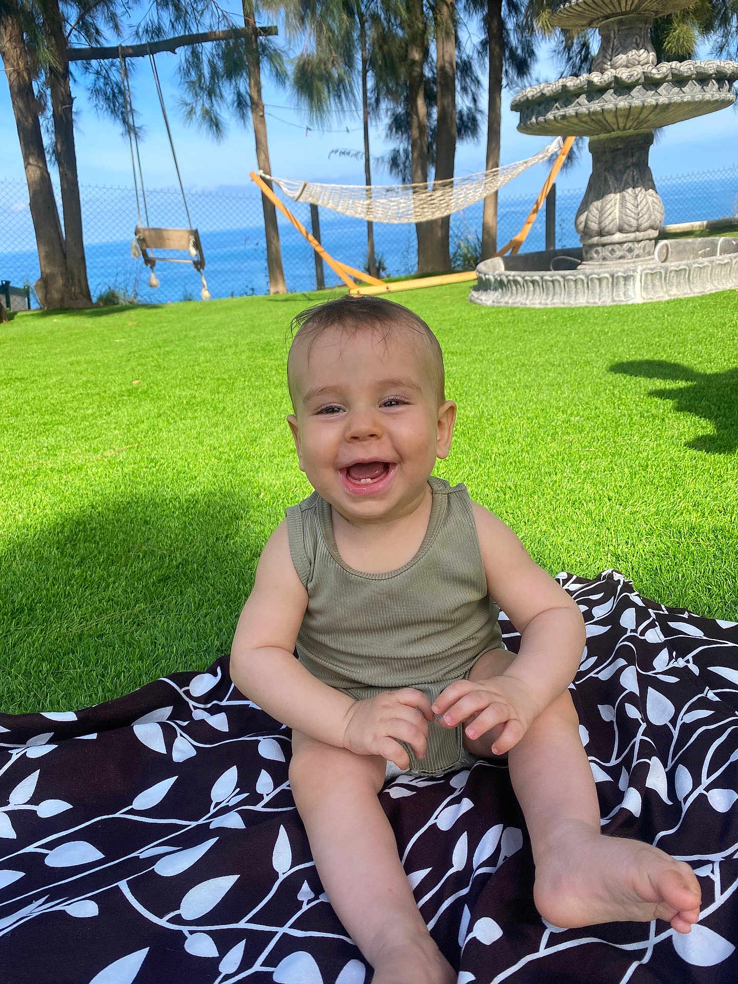 Ilyan participe au concours pour gagner de l'argent avec cette photo : baby, child, grass, outdoor, smiling, happy, blanket, nature, trees, blue_sky, sunlight, infant, playful, summer, portrait, grass_field, sitting, clothing, fence, fountain