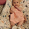 baby, infant, child, sitting, clothing, pink_dress, baby_chair, patterned, fabric, footwear, indoor, cute, hand_in_mouth, person, face, young_child, soft_lighting, bedroom, comfort, curious