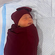 Rose is registered to the contest to win money with this photo: newborn, baby, sleeping, swaddled, infant, portrait, face, closed_eyes, indoors, changing_pad, blanket, burgundy, hat, peaceful, resting, photograph, phone_screen, gallery_interface, quilted_surface, baby_care