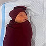 newborn, baby, sleeping, swaddled, infant, portrait, face, closed_eyes, indoors, changing_pad, blanket, burgundy, hat, peaceful, resting, photograph, phone_screen, gallery_interface, quilted_surface, baby_care