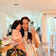 Muriel Kim is registered to the contest to win money with this photo: woman, toddler, child, indoor, living_room, mirror_selfie, smartphone, smiling, dog, white_dog, furniture, curtains, window, tiled_floor, handbag, plant, modern_interior, person, holding, home