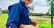 Leonardo is registered to the contest to win money with this photo: child, toddler, grass, outdoor, blue_shirt, tan_pants, toy, pink_cup, plush_toy, sky, clouds, daylight, greenery, hand, face, curious, sitting, nature, playtime, young_boy