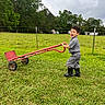 boots, boy, casual_clothing, child, cloudy_sky, farm, fence, field, grass, greenery, happy, nature, outdoor, play, rural, smile, toy, trees, wheelbarrow, young