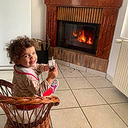 Lana participe au concours pour gagner de l'argent avec cette photo : toddler, child, smiling, wicker_chair, curly_hair, fireplace, indoor, tile_floor, holding_object, wall_decor, radiator, cozy, home, happy, seated, furniture, clothing, toy, warm, person
