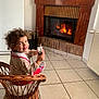 toddler, child, smiling, wicker_chair, curly_hair, fireplace, indoor, tile_floor, holding_object, wall_decor, radiator, cozy, home, happy, seated, furniture, clothing, toy, warm, person
