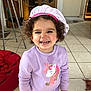 child, toddler, smiling, curly_hair, chef_hat, purple_clothing, unicorn, indoor, kitchen, tile_floor, wooden_doors, red_bean_bag, happy, cute, person, playful, young, home, casual, standing