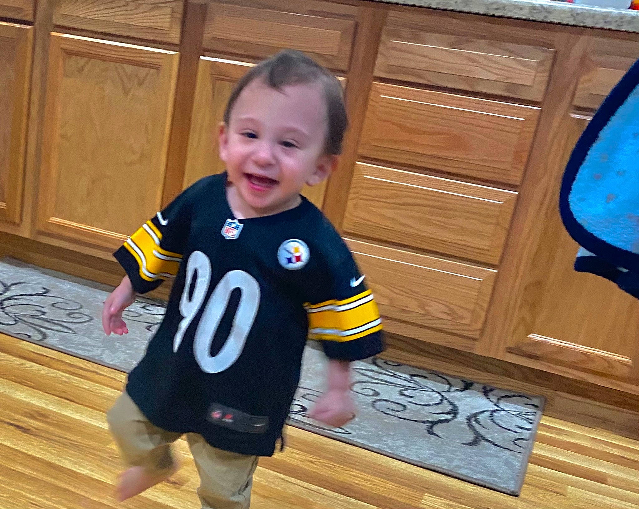 Benjamin is registered to the contest to win money with this photo: baby, baby_toddler_clothing, facial_expression, floor, flooring, fun, hairstyle, happy, hardwood, jersey, joy, person, sleeve, smile, sportswear, standing, t_shirt, television, toddler, varnish