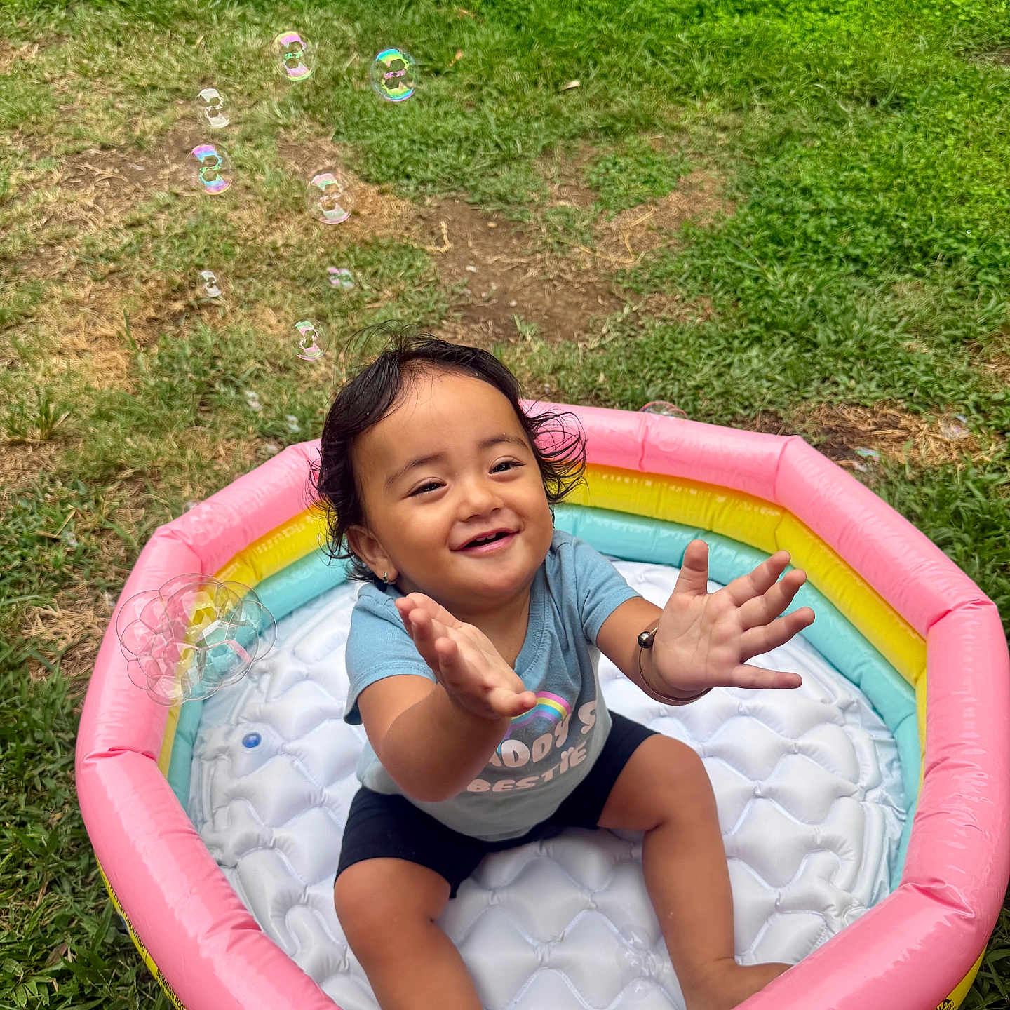 Tania is registered to the contest to win money with this photo: toddler, child, inflatable_pool, bubbles, smiling, outdoor, grass, play, happy, summer, water_toy, colorful, fun, baby, person, sitting, joyful, casual_clothing, nature, park