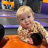 Angelina joined the competition — help win amazing prizes! toddler, child, indoor, trampoline_park, orange_surface, purple_padding, black_mesh_container, curious_expression, floral_shirt, red_bow, playful, person, baby, head, face, hand, background, colorful, indoor_play_area, young_child