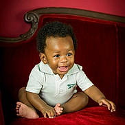 Kayceon is registered to the contest to win money with this photo: baby, child, smiling, sitting, white_shirt, polo_shirt, red_sofa, velvet_sofa, indoor, cute, happy, portrait, infant, person, furniture, curly_hair, young_child, bright_eyes, barefoot, cozy