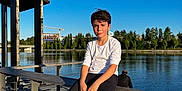 Mauricio a rejoint le concours — aidez-le/la à gagner de superbes lots ! boy, child, river, water, outdoor, daylight, blue_sky, trees, bench, railing, casual_clothing, white_shirt, black_pants, sneakers, adolescent, sunlight, reflection, relaxing, nature, park