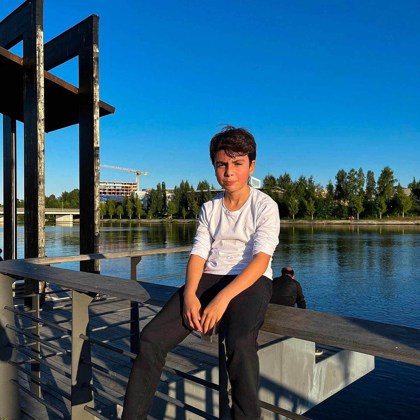 Mauricio a rejoint le concours — aidez-le/la à gagner de superbes lots ! adolescent, bench, black_pants, blue_sky, boy, casual_clothing, child, daylight, nature, outdoor, park, railing, reflection, relaxing, river, sneakers, sunlight, trees, water, white_shirt