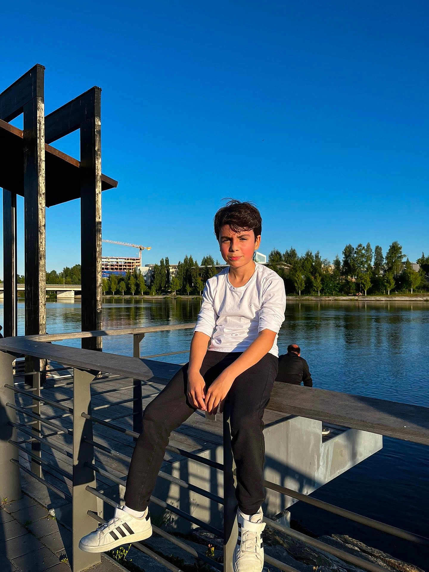 Mauricio a rejoint le concours — aidez-le/la à gagner de superbes lots ! boy, child, river, water, outdoor, daylight, blue_sky, trees, bench, railing, casual_clothing, white_shirt, black_pants, sneakers, adolescent, sunlight, reflection, relaxing, nature, park