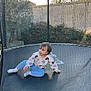 child, toddler, trampoline, safety_net, mesh, backyard, fence, grass, trees, sweater, leggings, socks, sitting, playing, outdoor, portrait, sunlight, garden_wall, netting, short_hair