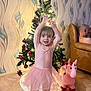toddler, child, pink_tutu, christmas_tree, holiday_lights, toy_unicorn, living_room, brown_couch, wallpaper, floor_tiles, smiling, happy, indoor, holiday_decor, person, plush_toy, festive, cute, barefoot, home