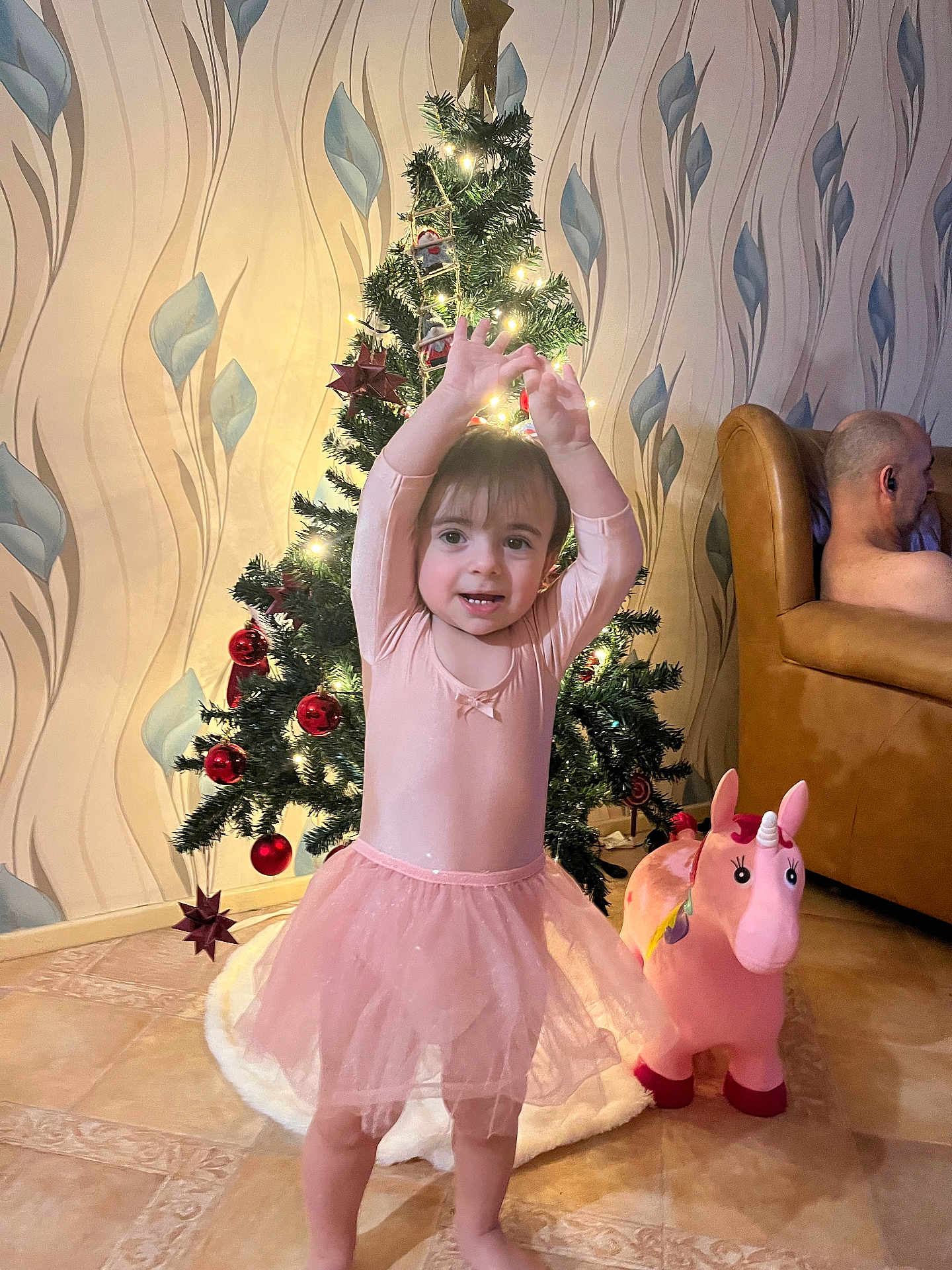 Pereira Marcia a rejoint le concours — aidez-le/la à gagner de superbes lots ! toddler, child, pink_tutu, christmas_tree, holiday_lights, toy_unicorn, living_room, brown_couch, wallpaper, floor_tiles, smiling, happy, indoor, holiday_decor, person, plush_toy, festive, cute, barefoot, home