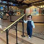 Pereira Marcia a rejoint le concours — aidez-le/la à gagner de superbes lots ! child, toddler, museum, exhibit, dinosaur_skeleton, display_case, handrail, railing, stairs, tiled_floor, overalls, smile, portrait, indoor, family, adult, glass, lighting, decor, visitor