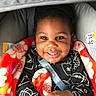 toddler, child, smiling, blanket, car_seat, seatbelt, black_clothing, patterned_clothing, indoors, happy, face, hands, cute, baby, portrait, infant, warm, colorful, cozy, young_child