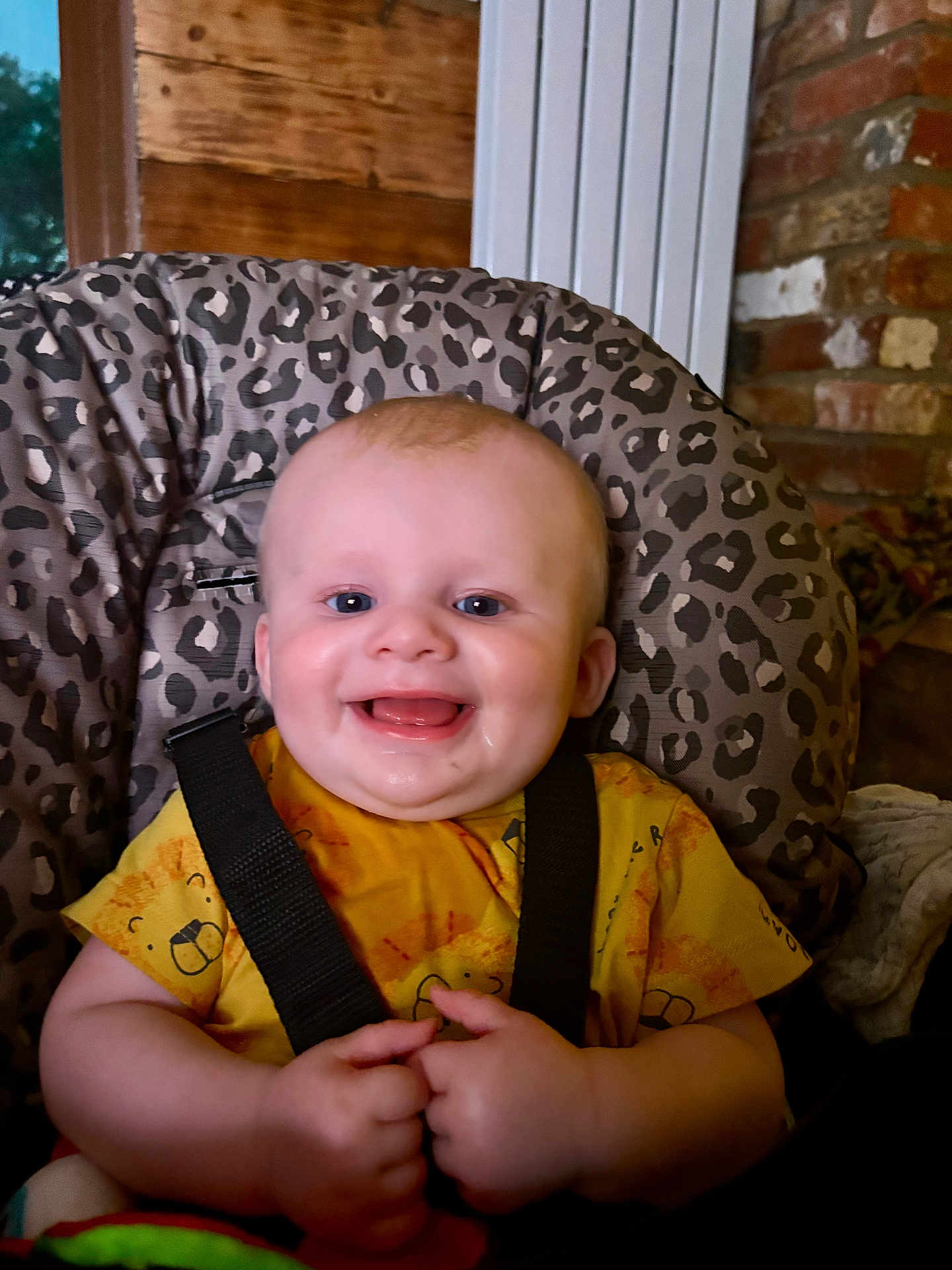 Finlay is registered to the contest to win money with this photo: baby, child, smiling, happy, infant, yellow_shirt, animal_print, high_chair, leopard_print, indoor, seat_belt, wooden_background, brick_wall, hands, face, cute, portrait, baby_clothing, human, young_child