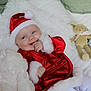 baby, santa_hat, red_clothing, white_blanket, plush_toy, gift_box, holiday, festive, smiling, cute, infant, cozy, soft_toy, christmas, lying_down, happy, white_tights, indoors, child, celebration