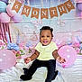 baby, child, smiling, balloon, banner, 6_months, half_year, celebration, portrait, sitting, yellow_top, black_pants, curly_hair, beads, decorations, floral_backdrop, butterflies, pastel_colors, happy, indoor