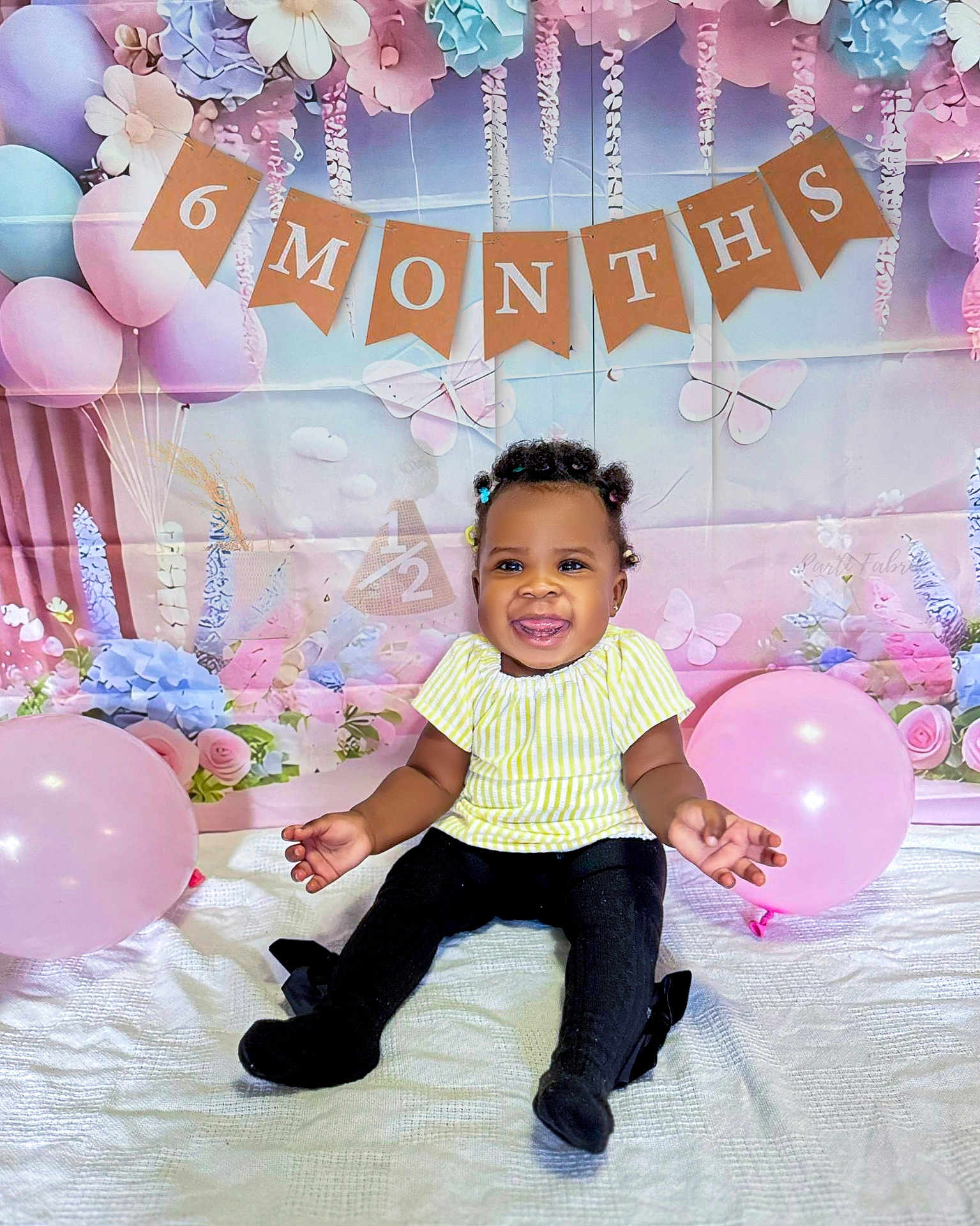 Zoey joined the competition — help win amazing prizes! baby, child, smiling, balloon, banner, 6_months, half_year, celebration, portrait, sitting, yellow_top, black_pants, curly_hair, beads, decorations, floral_backdrop, butterflies, pastel_colors, happy, indoor