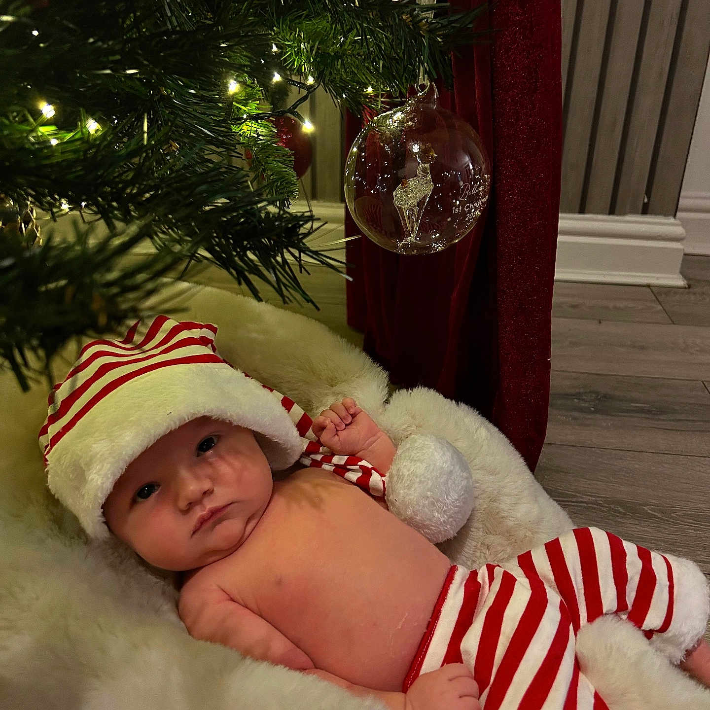 Bellamy is registered to the contest to win money with this photo: baby, blanket, celebration, christmas, cozy, cute, decorations, festive, floor, hat, holiday, indoors, infant, lighting, ornament, pom_pom, red_and_white, striped_clothing, tree, wooden_floor