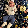 baby, smiling, mittens, headband, bow, milestone_card, blanket, floral_pattern, pants, booties, infant, cute, lying_down, happy, indoors, soft_texture, child, clothing, person, portrait