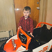 Karson joined the competition — help win amazing prizes! child, boy, toy_car, indoor, red_shirt, checkered_shirt, steering_wheel, wooden_door, paneled_wall, floor, head, face, person, vehicle, play, childhood, young, casual, furniture, room
