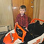 child, boy, toy_car, indoor, red_shirt, checkered_shirt, steering_wheel, wooden_door, paneled_wall, floor, head, face, person, vehicle, play, childhood, young, casual, furniture, room